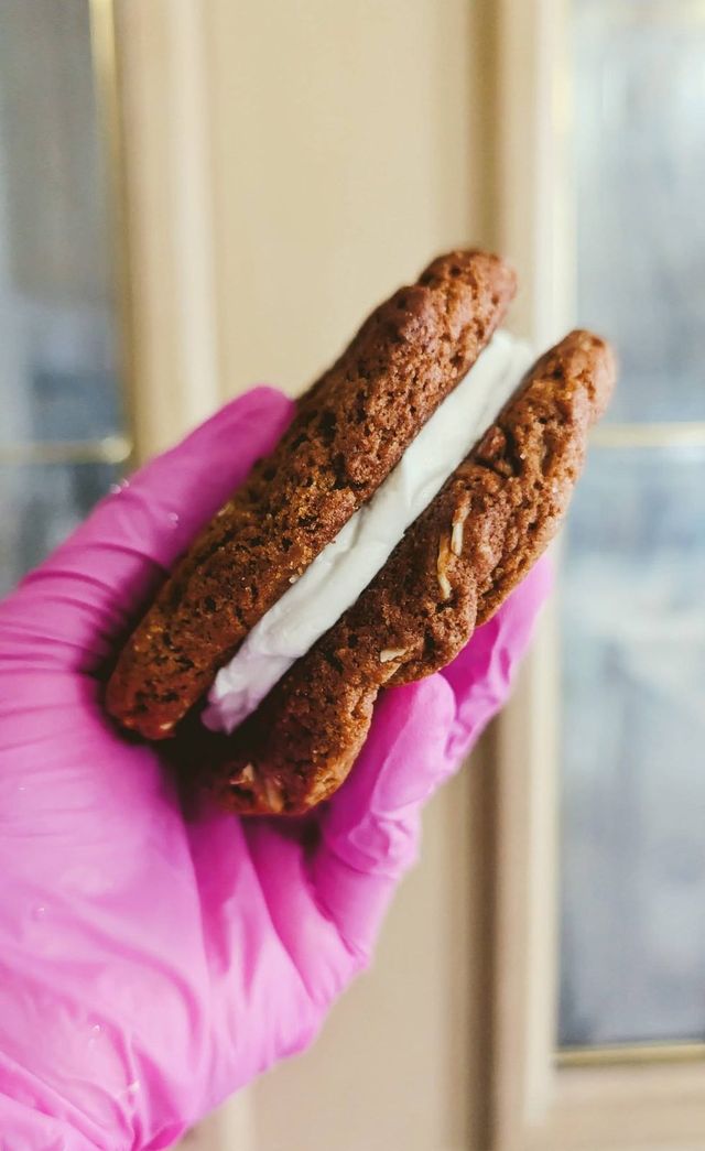 A pink-gloved hand holding a sandwich cookie filled with buttercream