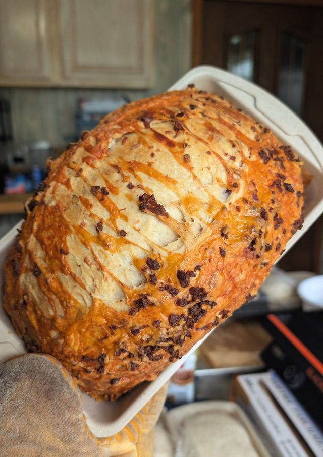 A golden loaf with melted cheese and dark bits on top, held up in its paper pan