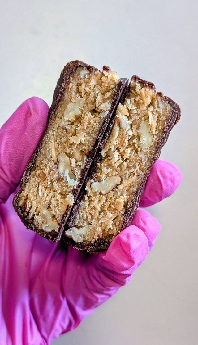 A pink-gloved hand holding a bar with a chocolate coating, cut in half to show a nutty filling